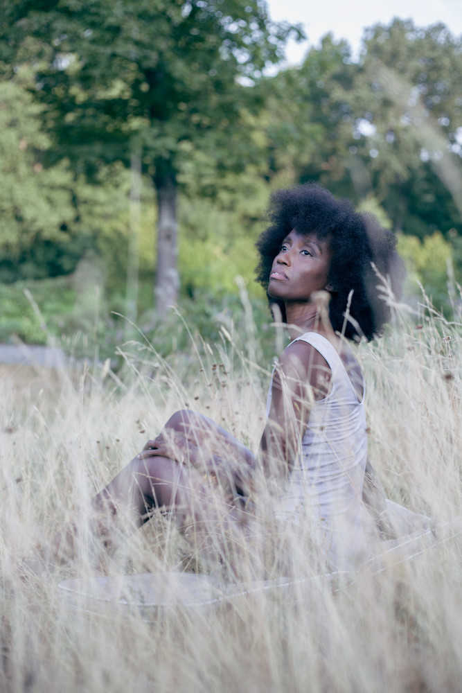 Eine junge schwarze Frau sitzt in einer Wiese. Sie trägt einen nachhaltigen Body in Lila