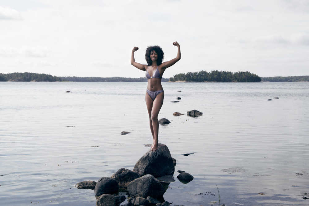 Eine junge schwarze Frau steht auf Steinen vor einem See in starker Pose. Sie trägt nachhaltige Unterwäsche aus Seide in Lila