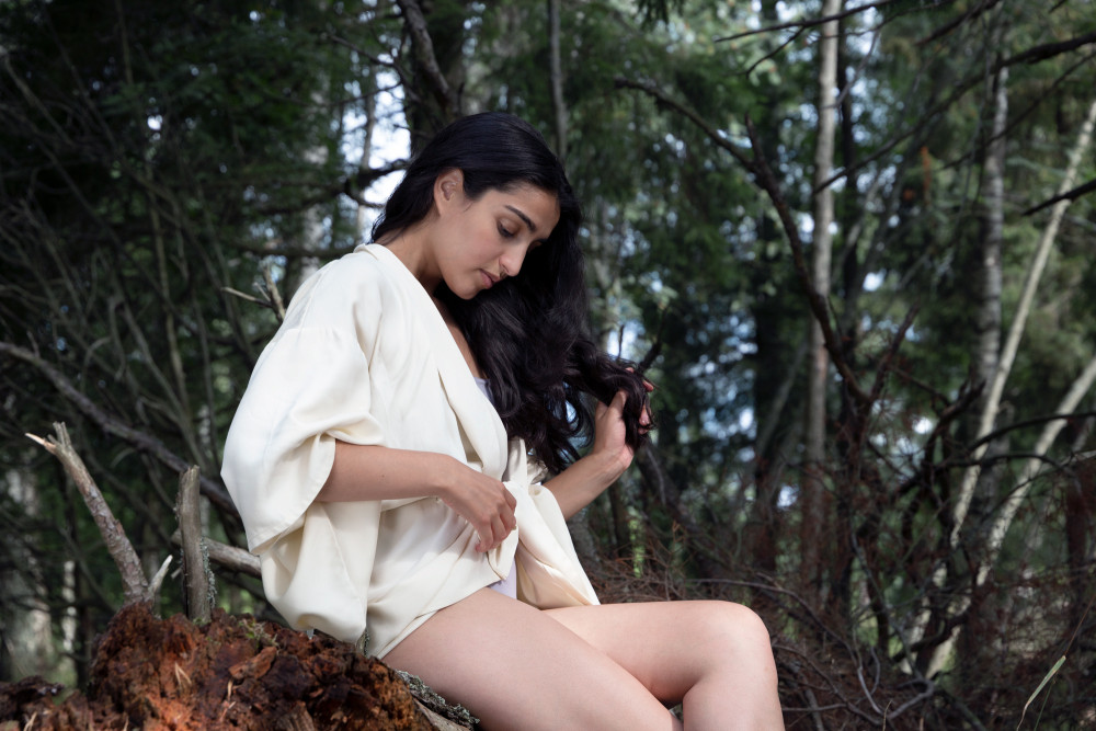 Eine junge Frau sitzt im Wald auf einem liegenden Baumstamm. Sie trägt einen nachhaltigen weißen Kimono aus Tencel.